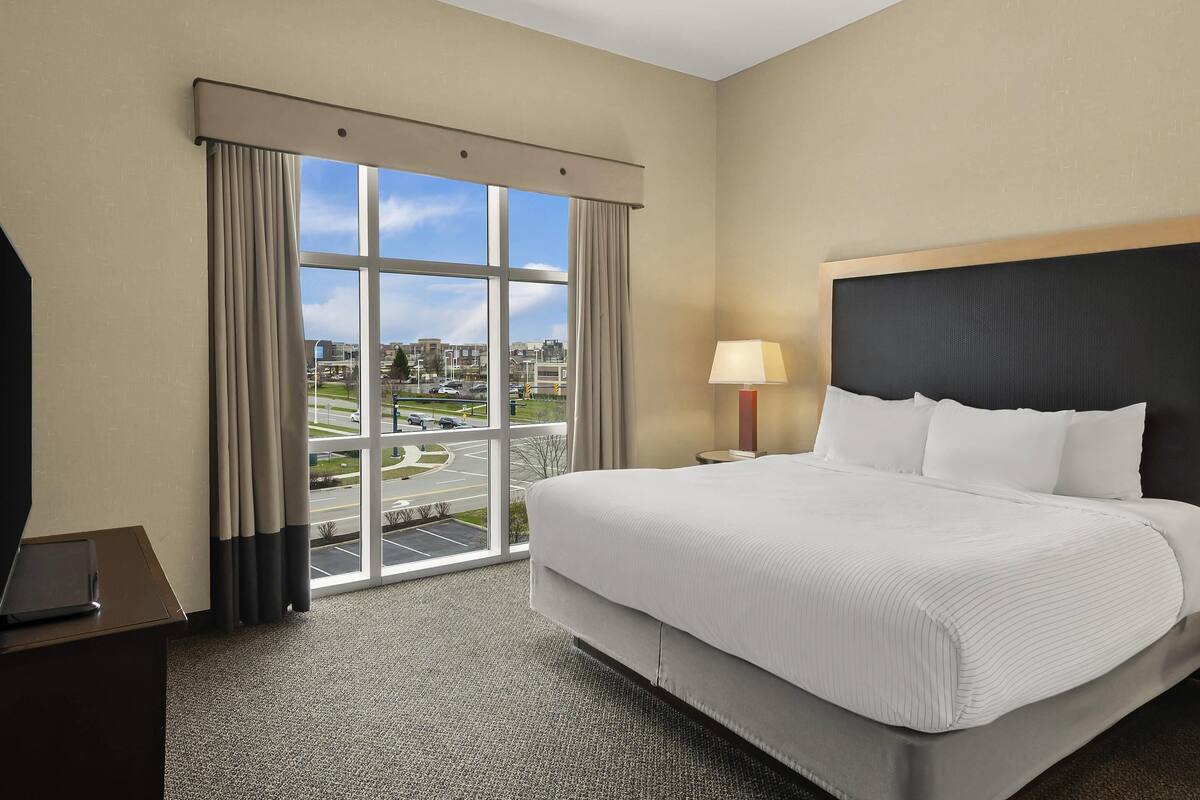 Ein Hotelzimmer mit einem großen Bett, einem Schreibtisch, einer Lampe und Blick auf die Stadt durch ein Fenster.