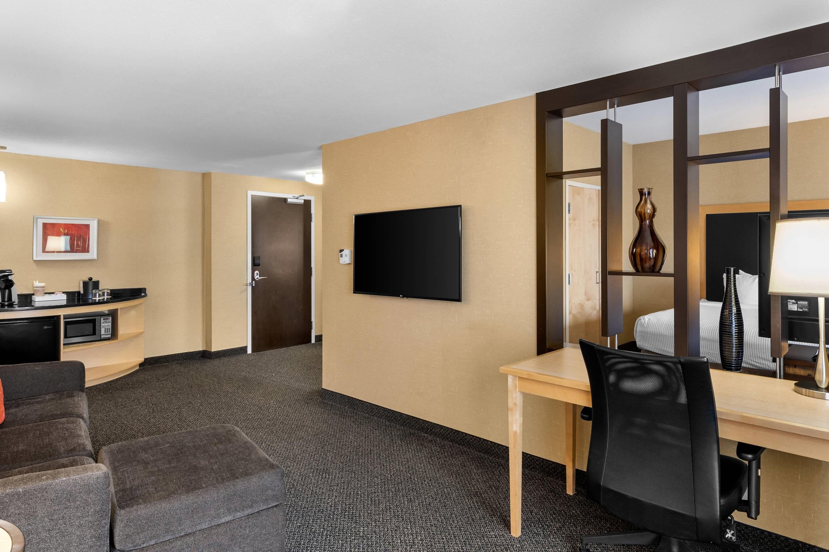 A hotel room with a desk, a television, a sofa, and a small kitchenette.
