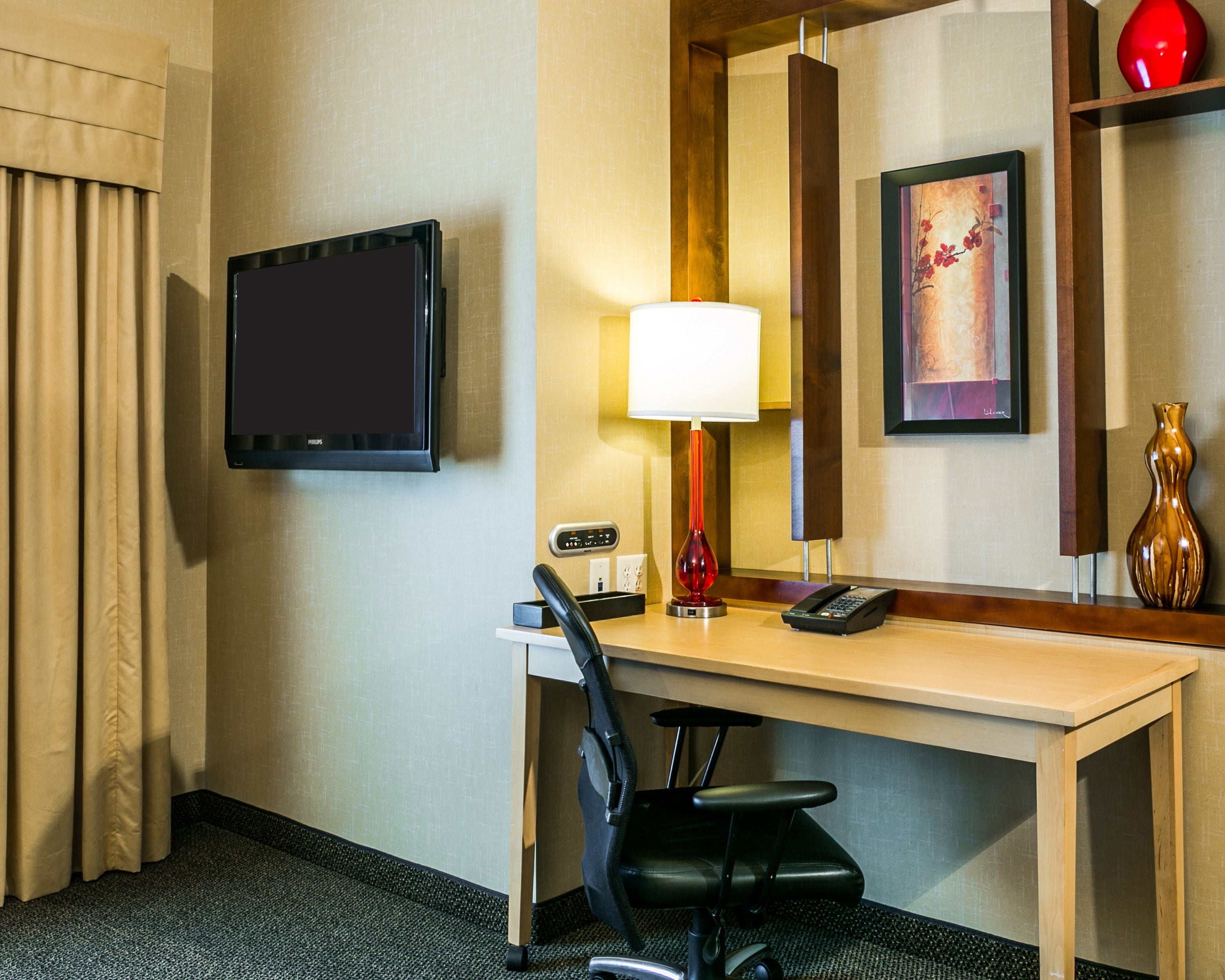 A hotel room with a desk, chair, lamp, phone, and artwork on the wall.