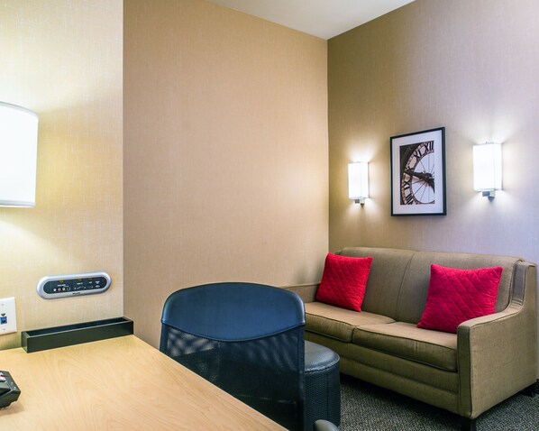 Habitación de hotel con un sofá color beige, dos almohadas rojas, una silla negra, una mesa de madera, un panel de control Philips y un reloj en la pared.