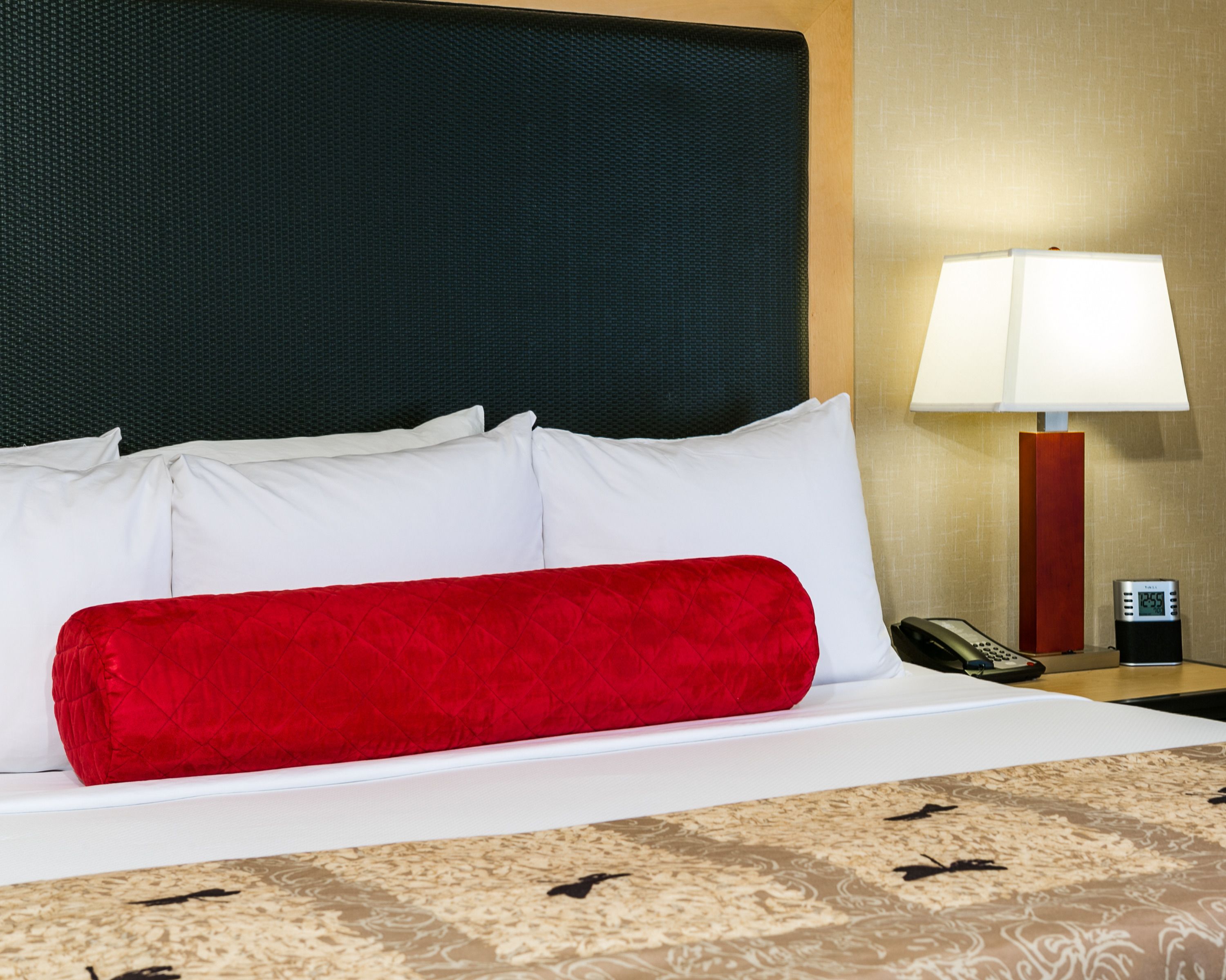 Una cama bien hecha con almohadas blancas y rojas, una colcha estampada, un cabecero negro, una lámpara de noche y una mesita de noche con un teléfono y un reloj digital.