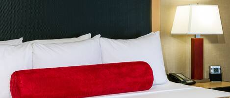 Una cama bien hecha con almohadas blancas y rojas, una colcha estampada, un cabecero negro, una lámpara de noche y una mesita de noche con un teléfono y un reloj digital.