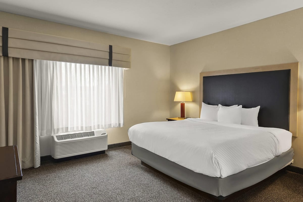 Ein Hotelzimmer mit einem großen Bett, einer Nachttischlampe, einem Fenster mit Vorhängen und einer Wand-Klimaanlage.