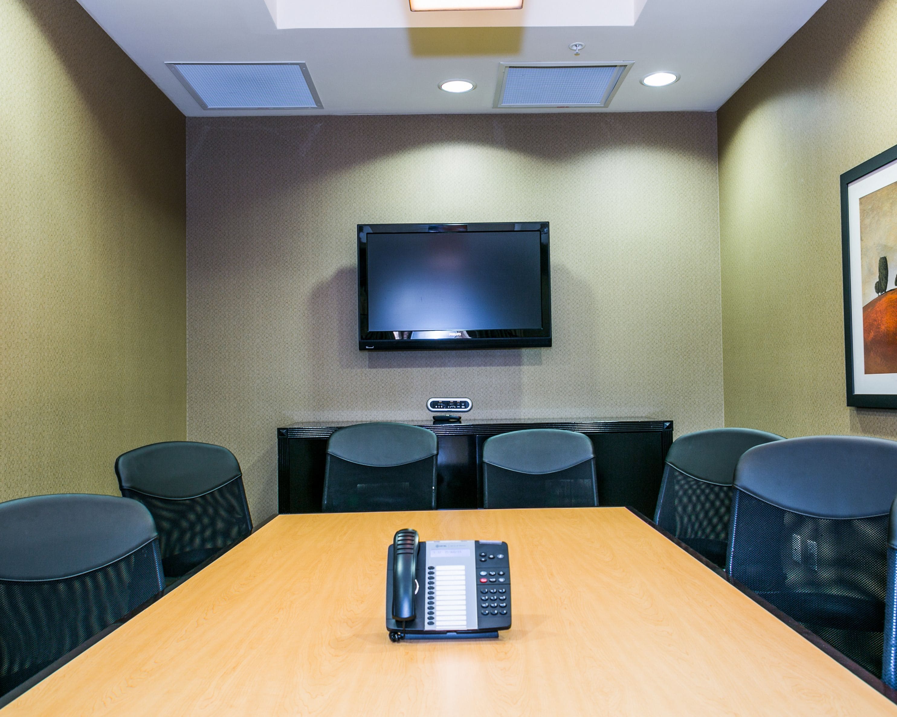A conference room with a wooden table, black chairs, a mounted flat-screen TV, and a telephone on the table.