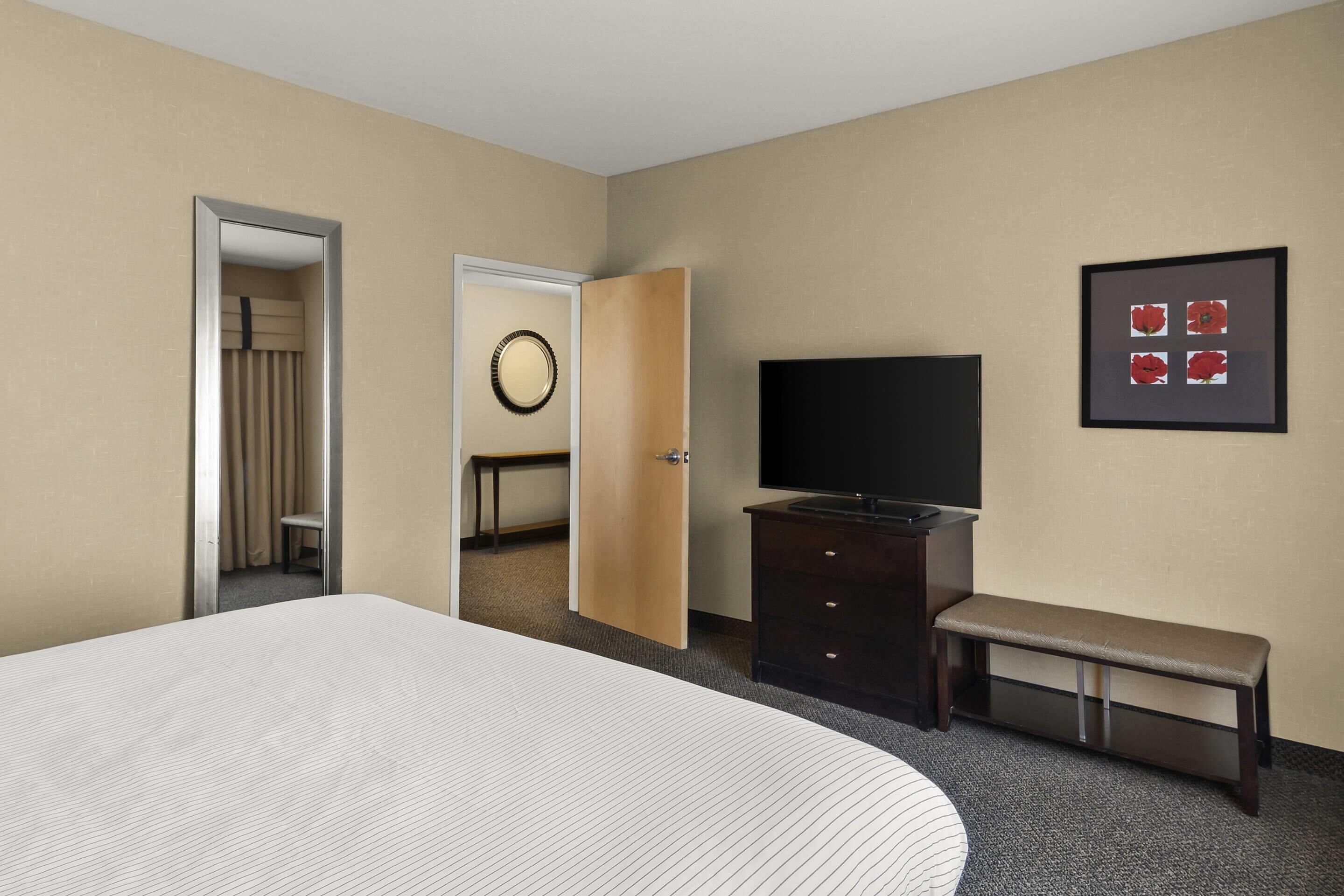 A hotel room with a large bed, a TV, a bench, a dresser, and a door leading to another room.