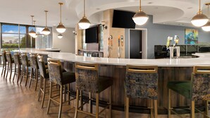 A modern bar area with a curved counter, multiple bar stools, pendant lighting, and a large window providing a view of the outdoors. - Cambria Hotel Columbus - Polaris (Columbus)