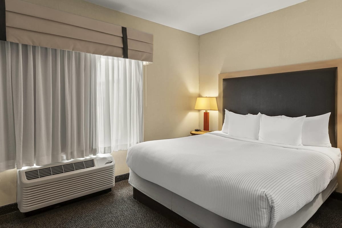 Ein Hotelzimmer mit einem großen Bett, einer Nachttischlampe, einem Fenster mit Vorhängen und einer Klimaanlage.