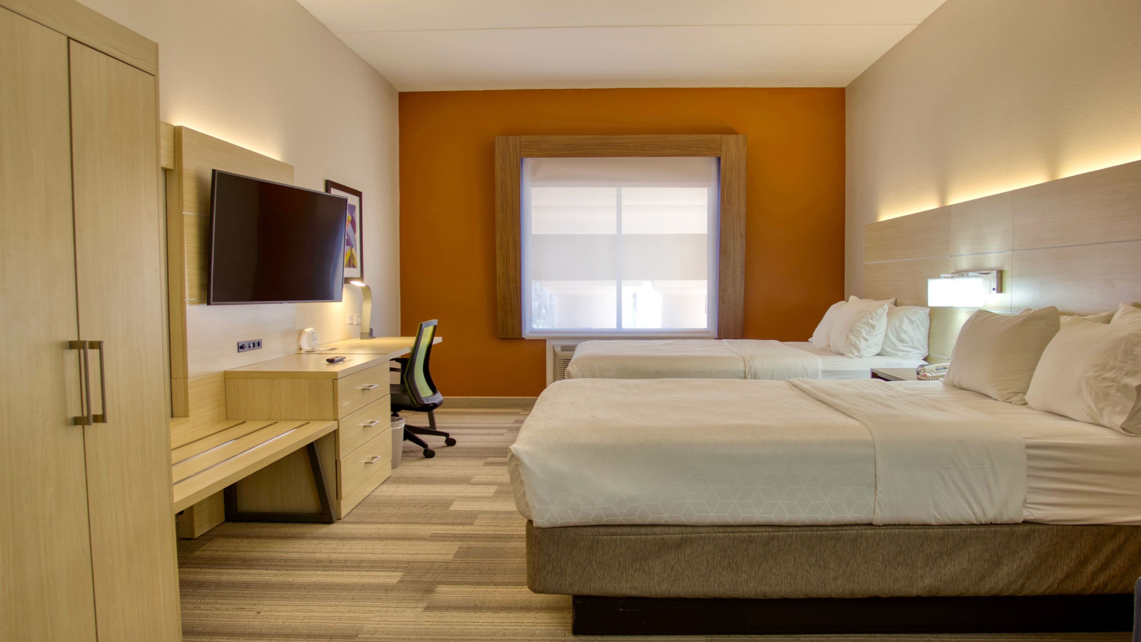Quarto de hotel com cama, escrivaninha, cadeira, televisão e janela com persianas.