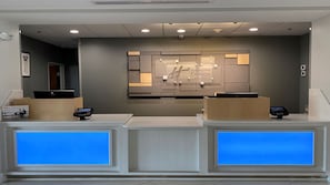 A hotel reception desk with two computer stations, a wall-mounted sign, and a blue-lit window. - Holiday Inn Express Hotel & Suites BYRAM by IHG (Byram)
