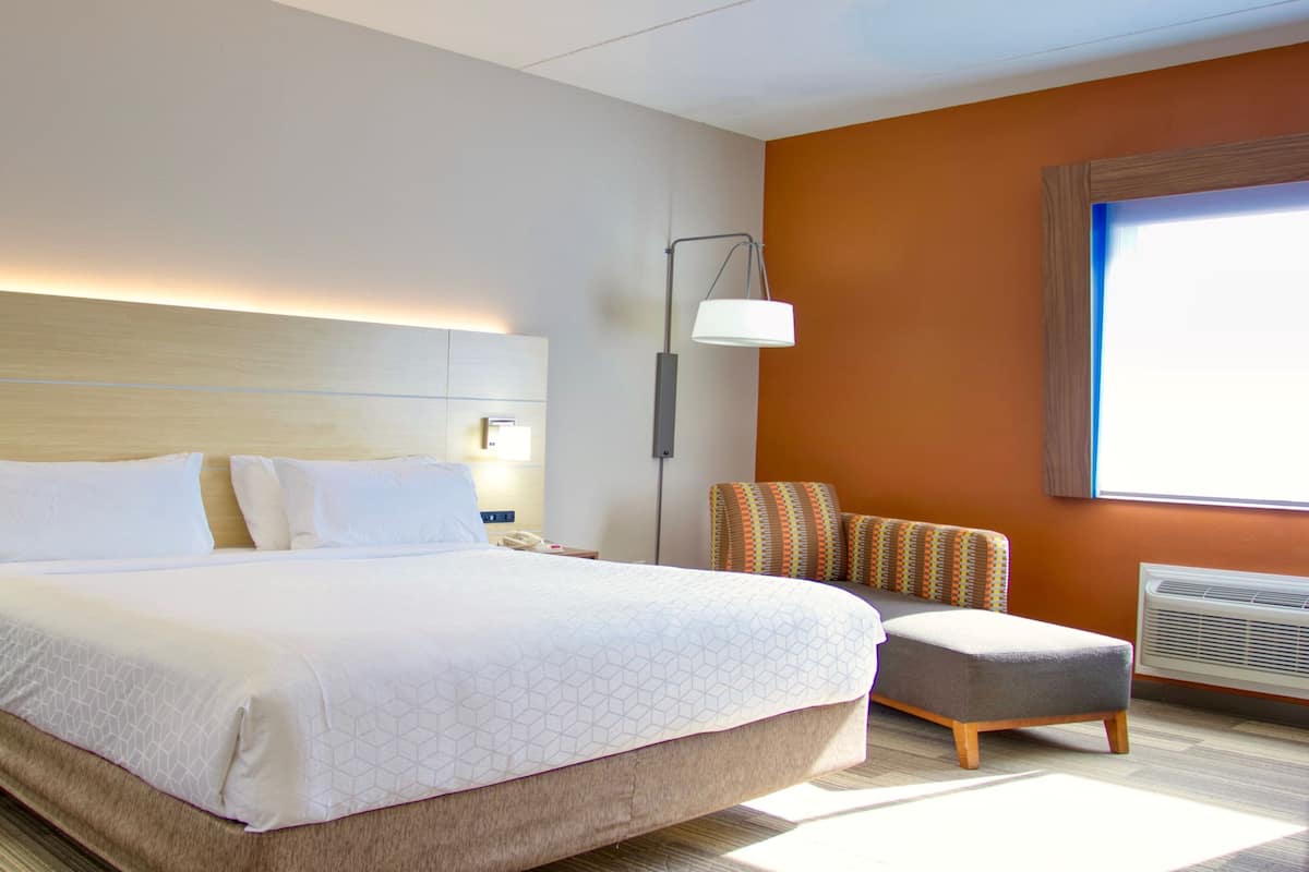 Une chambre d’hôtel avec un grand lit, une table de chevet avec une lampe, un pouf et une fenêtre avec des stores.