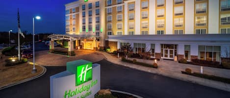 Un hôtel Holiday Inn & Suites, reconnaissable à son grand panneau, à ses nombreux étages et à son entrée bien éclairée.