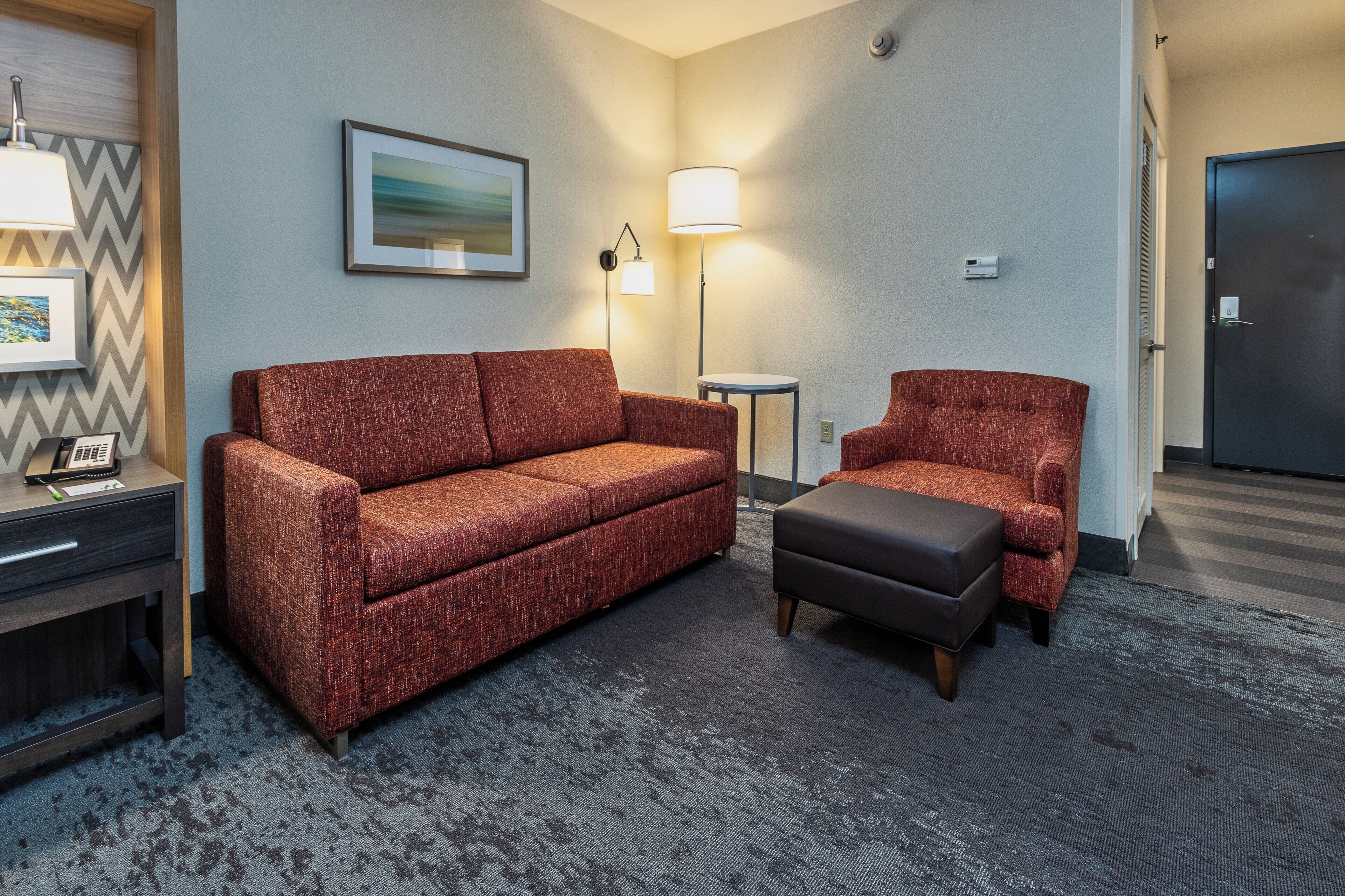A hotel room with a sofa, chair, and ottoman, a desk with a phone, and two wall-mounted lamps.