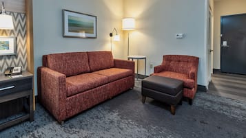 A hotel room with a sofa, chair, and ottoman, a desk with a phone, and two wall-mounted lamps.