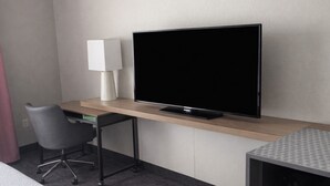 Une chambre d’hôtel avec un téléviseur à écran plat posé sur un bureau en bois, une chaise moderne et une petite table recouverte d’une nappe à motifs.