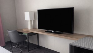 Une chambre d’hôtel avec un téléviseur à écran plat posé sur un bureau en bois, une chaise moderne et une petite table recouverte d’une nappe à motifs.