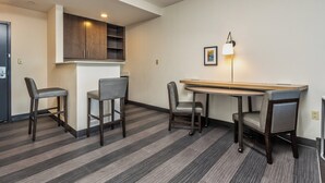 Une chambre d’hôtel moderne dotée d’un coin repas comprenant une longue table et quatre chaises, d’un coin cuisine avec des placards et d’une petite kitchenette.