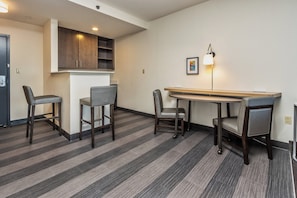 Une chambre d’hôtel moderne dotée d’un coin repas comprenant une longue table et quatre chaises, d’un coin cuisine avec des placards et d’une petite kitchenette.