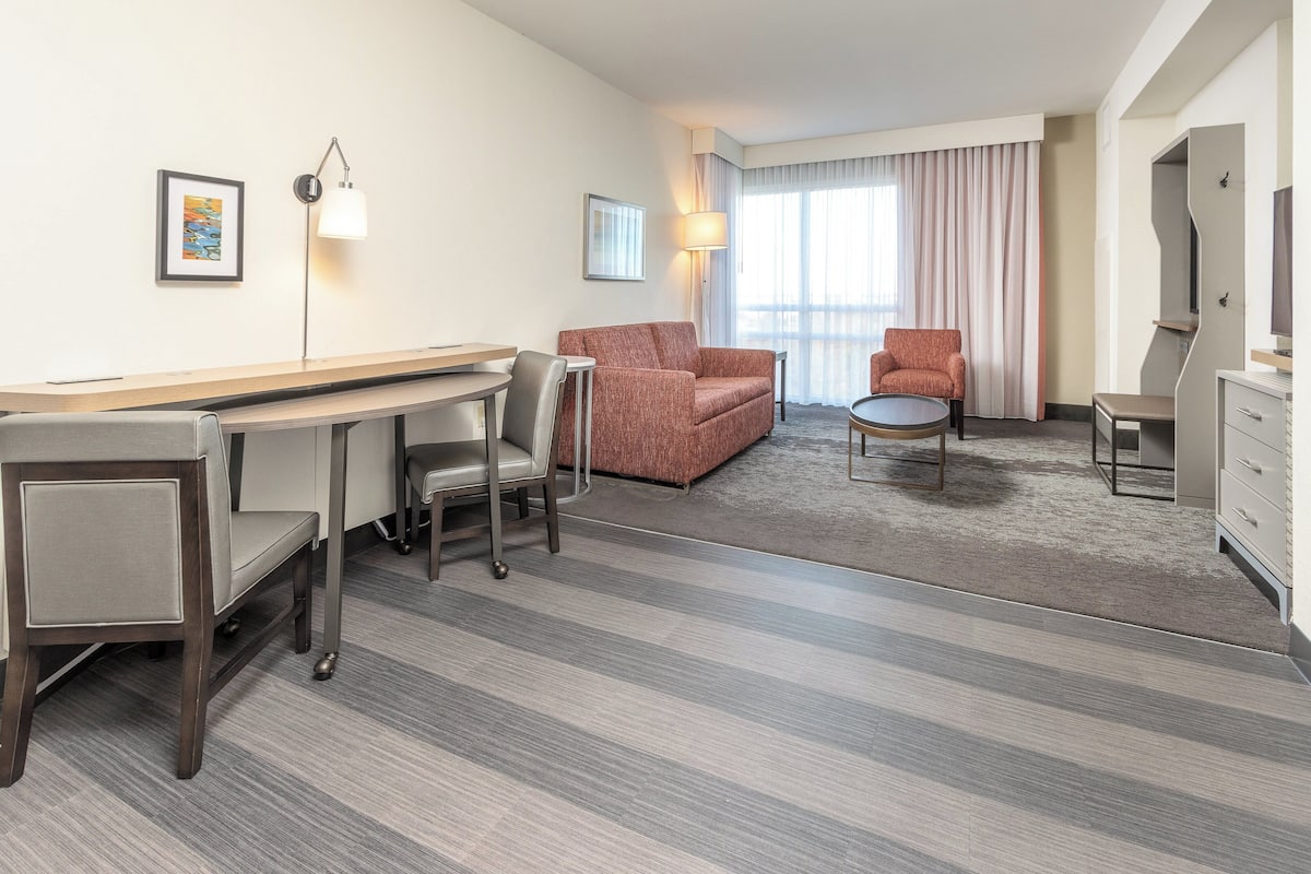 Hotelkamer met een bureau, twee fauteuils, een bank, een klein rond tafeltje, een televisie en een groot raam met gordijnen.