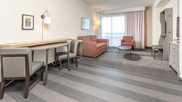 Habitación de hotel con escritorio, dos sillones, un sofá, una pequeña mesa redonda, televisor y una ventana grande con cortinas.