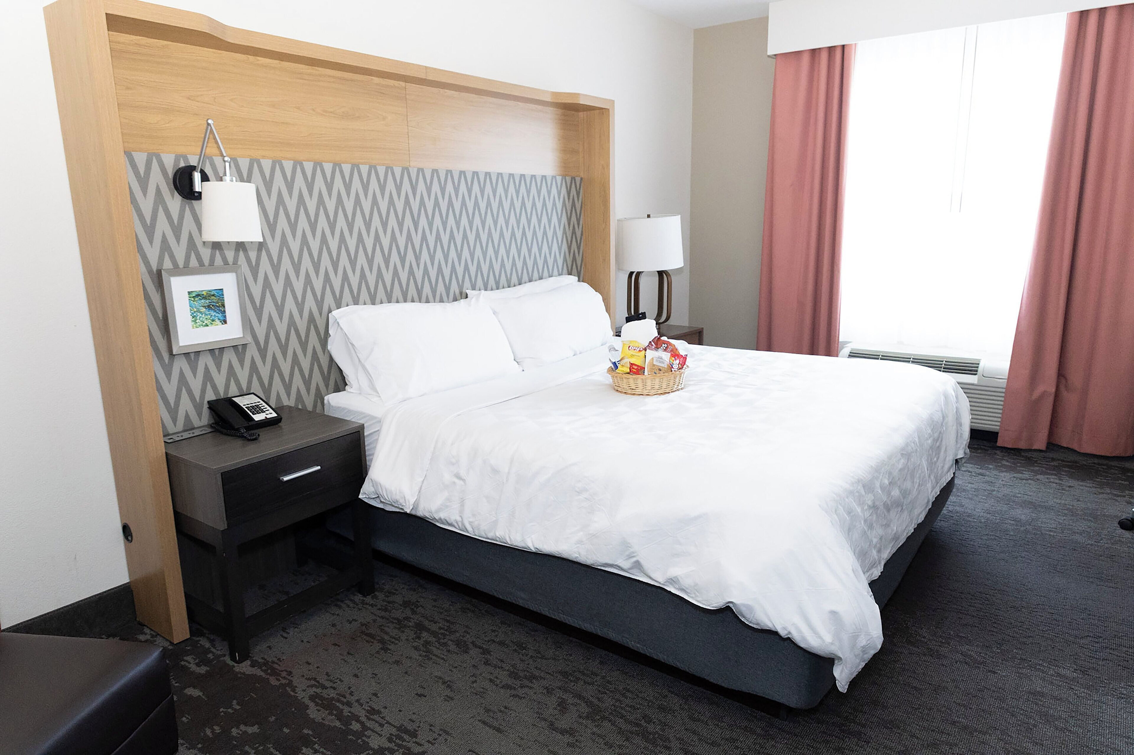 A hotel room with a bed, a bedside table with a phone, a lamp, a picture frame, and a basket of snacks.