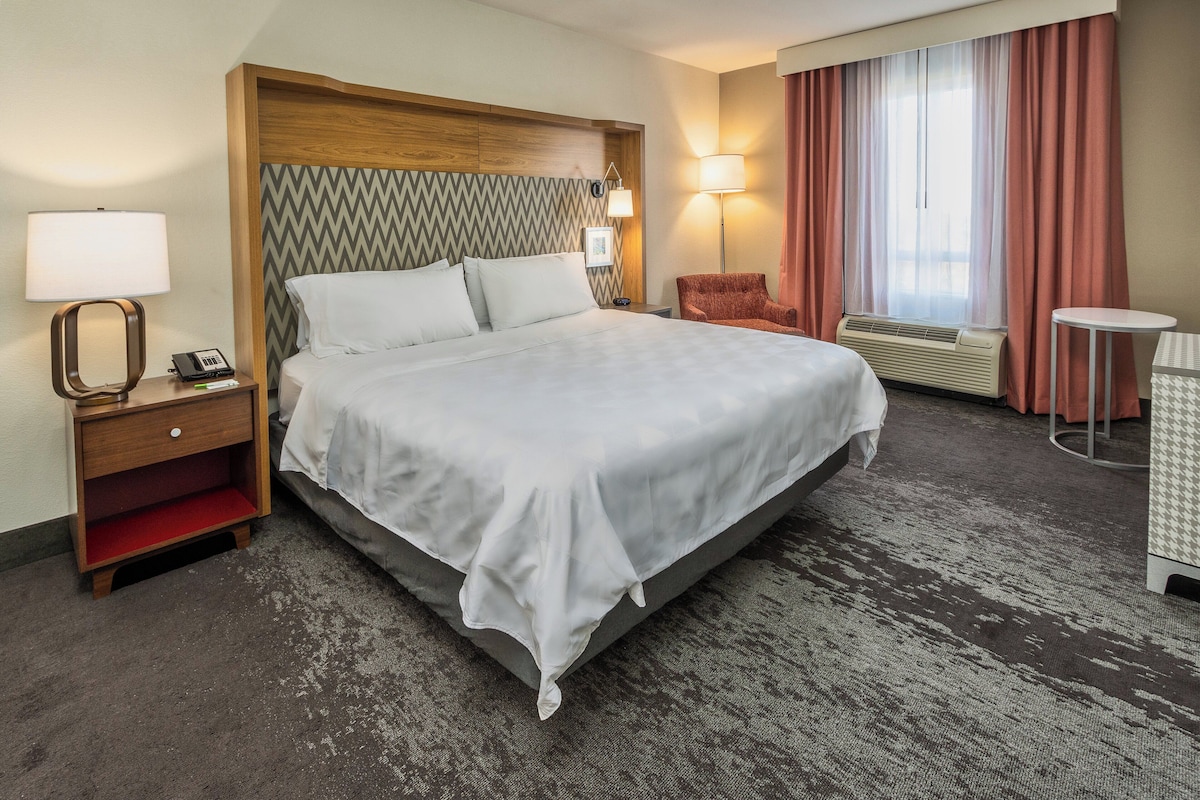 Une chambre d’hôtel comprenant un lit, une table de chevet avec une lampe, une chaise, une petite table et une fenêtre avec des rideaux.