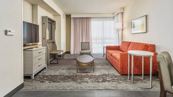 Habitación de hotel con sofá, una mesita redonda, televisor, escritorio con cajones, una silla y un ventanal con cortinas.