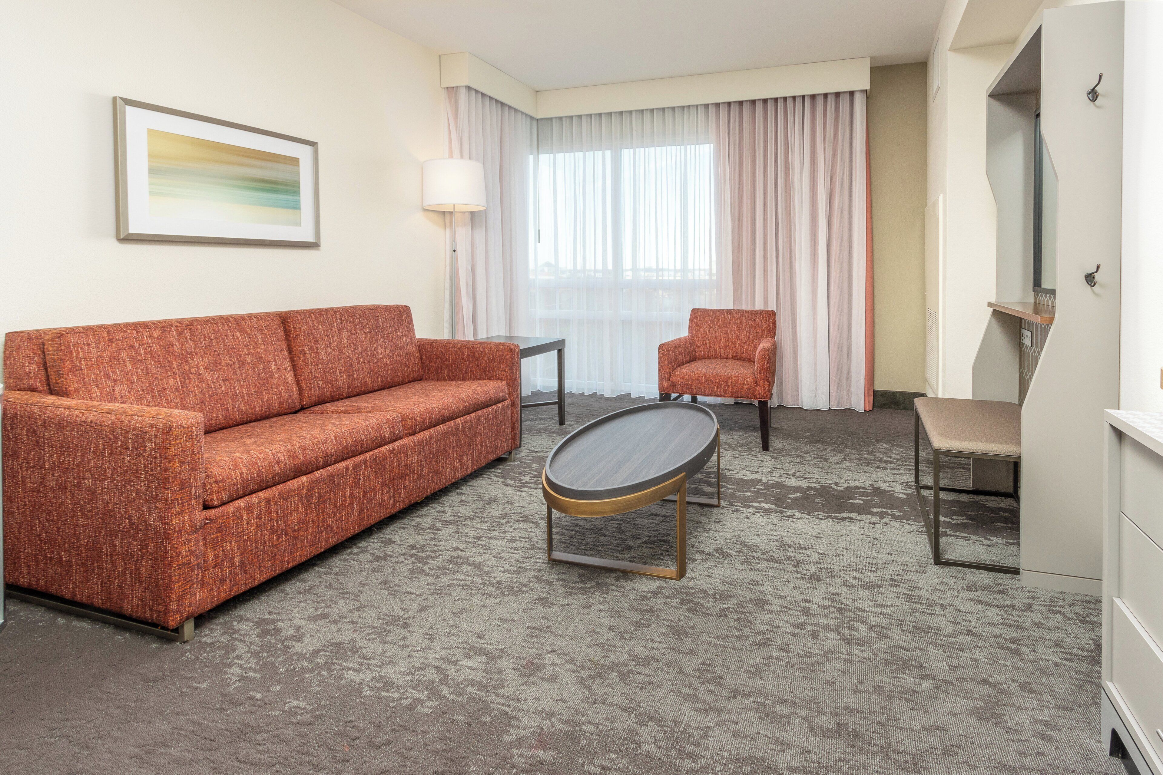 A hotel room with a sofa, armchair, small table, and a painting on the wall.