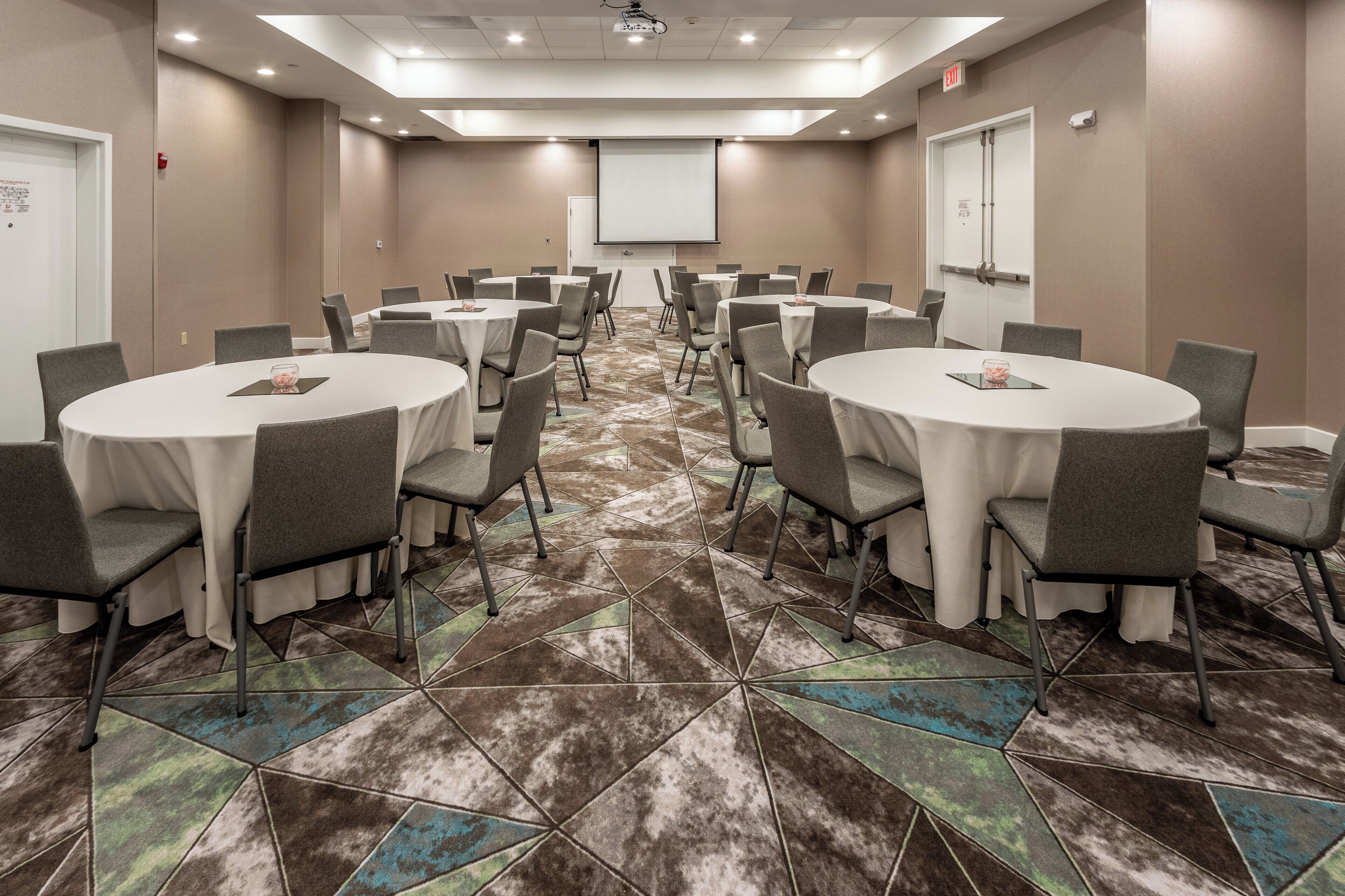 A conference room with multiple round tables and chairs, a projector screen, and a tiled floor.