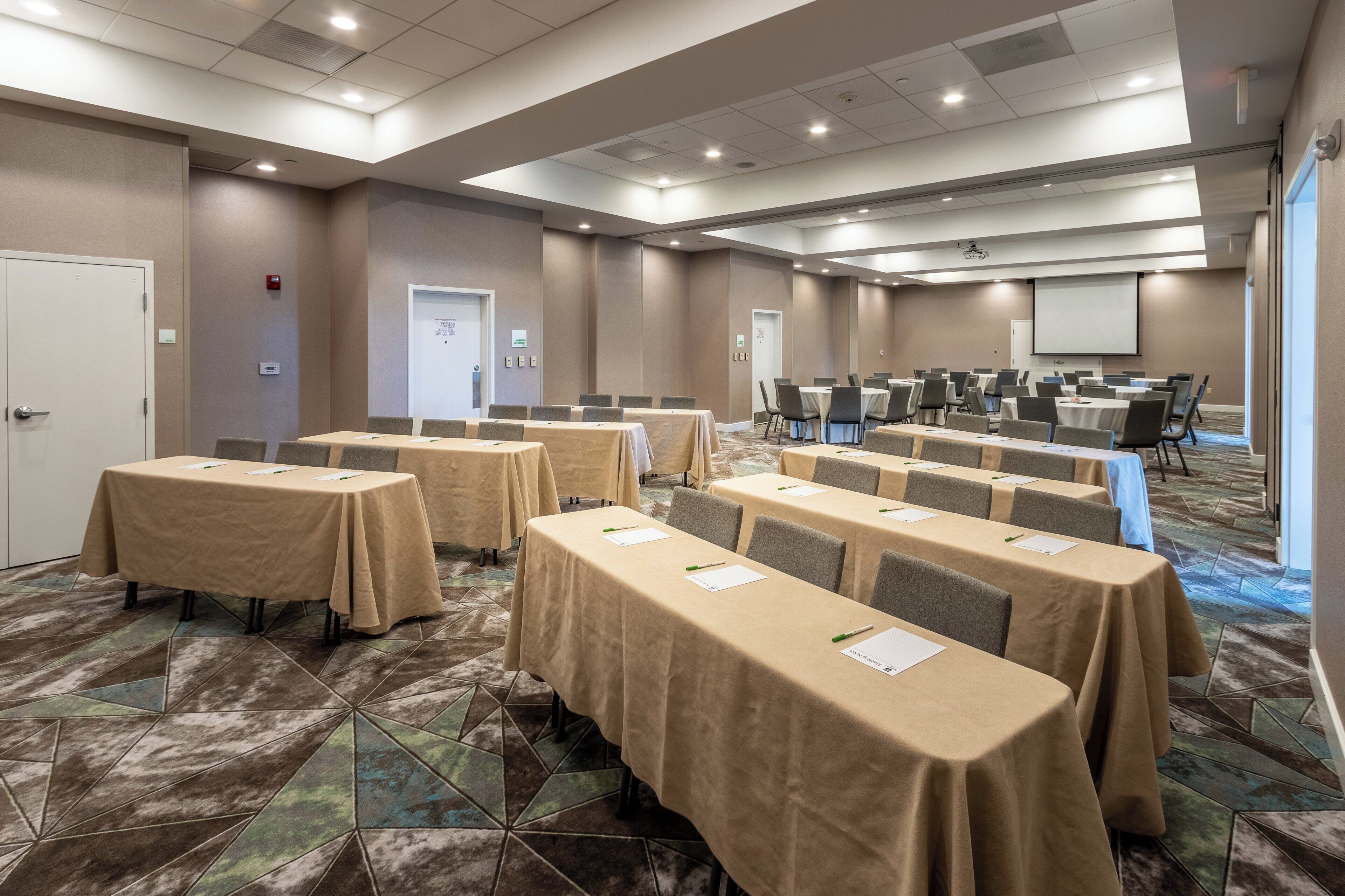 A conference room with multiple tables and chairs, equipped with nameplates and pens, and a projector screen at the front.