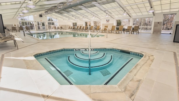 Une grande piscine intérieure dotée de plusieurs couloirs, entourée de chaises longues et surmontée d’une structure de toit abritée.