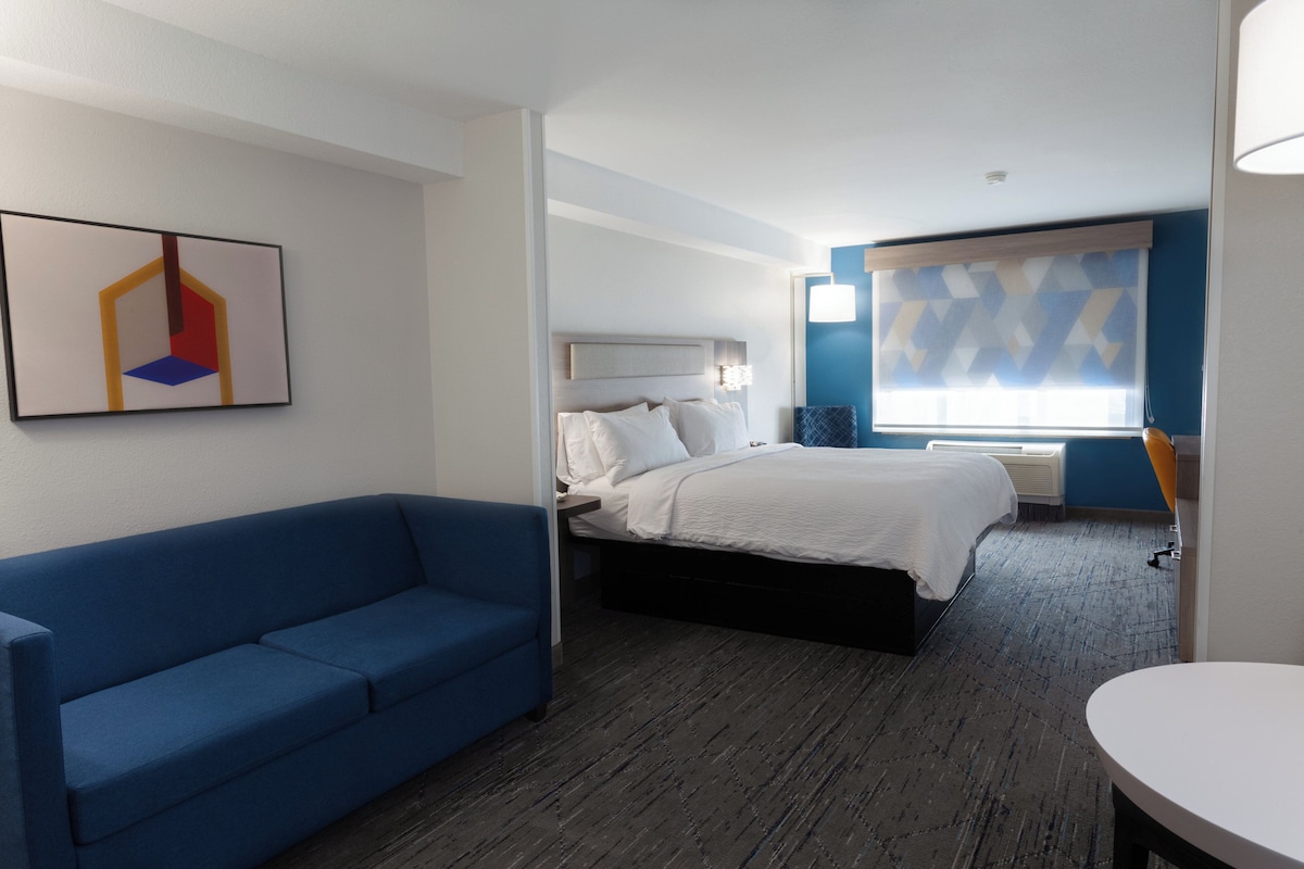 A hotel room with a bed, a blue sofa, a painting on the wall, and a window with blinds.