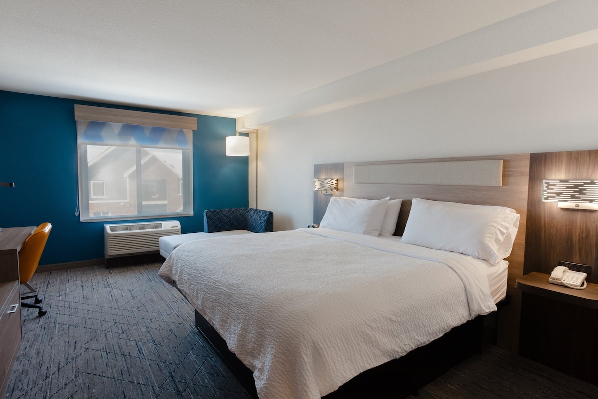 A hotel room with a large bed, a desk, a chair, a window, and a bedside table with a phone.