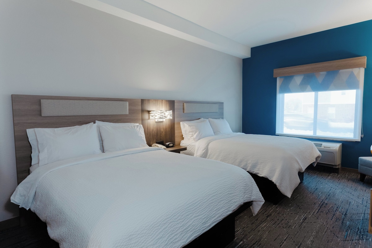 A hotel room with two beds, a wooden headboard, a nightstand, a window with a view, and a blue accent wall.