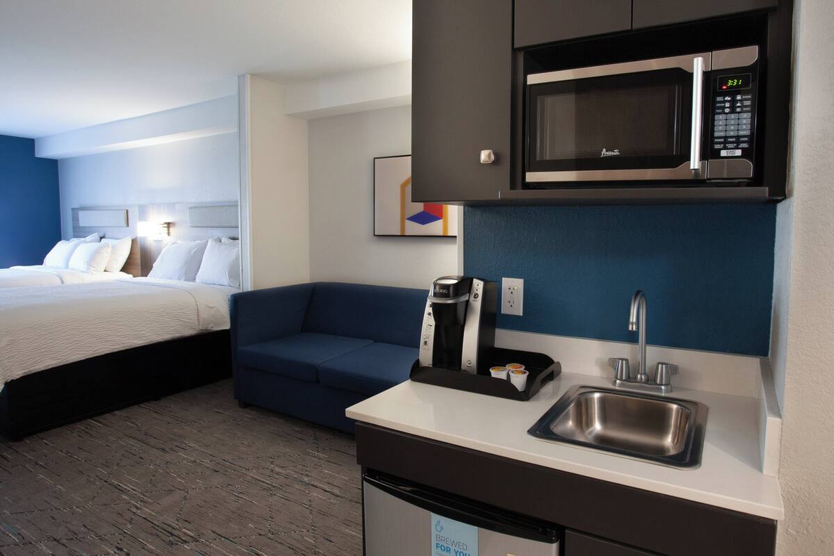 A hotel room with a bed, a sofa, a kitchenette with a microwave, a coffee maker, and a sink.