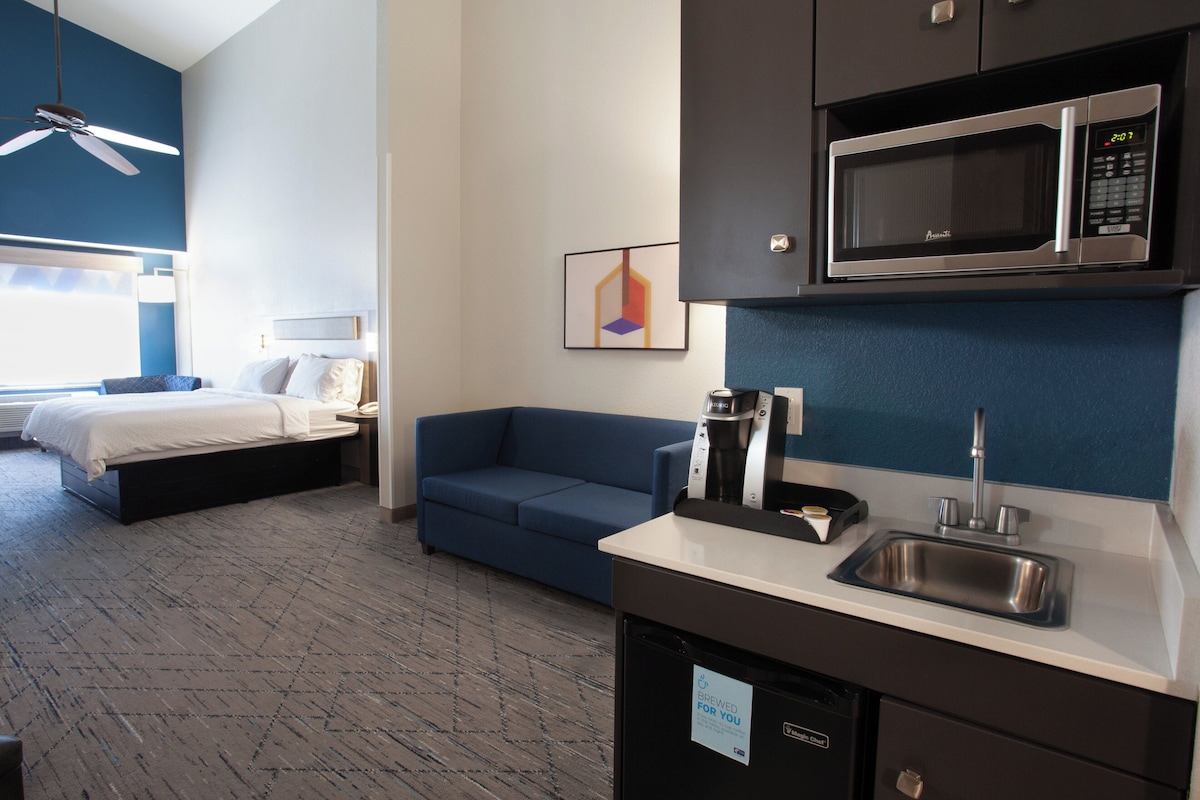 A hotel room with a bed, a sofa, a kitchenette with a microwave, a coffee maker, and a sink.