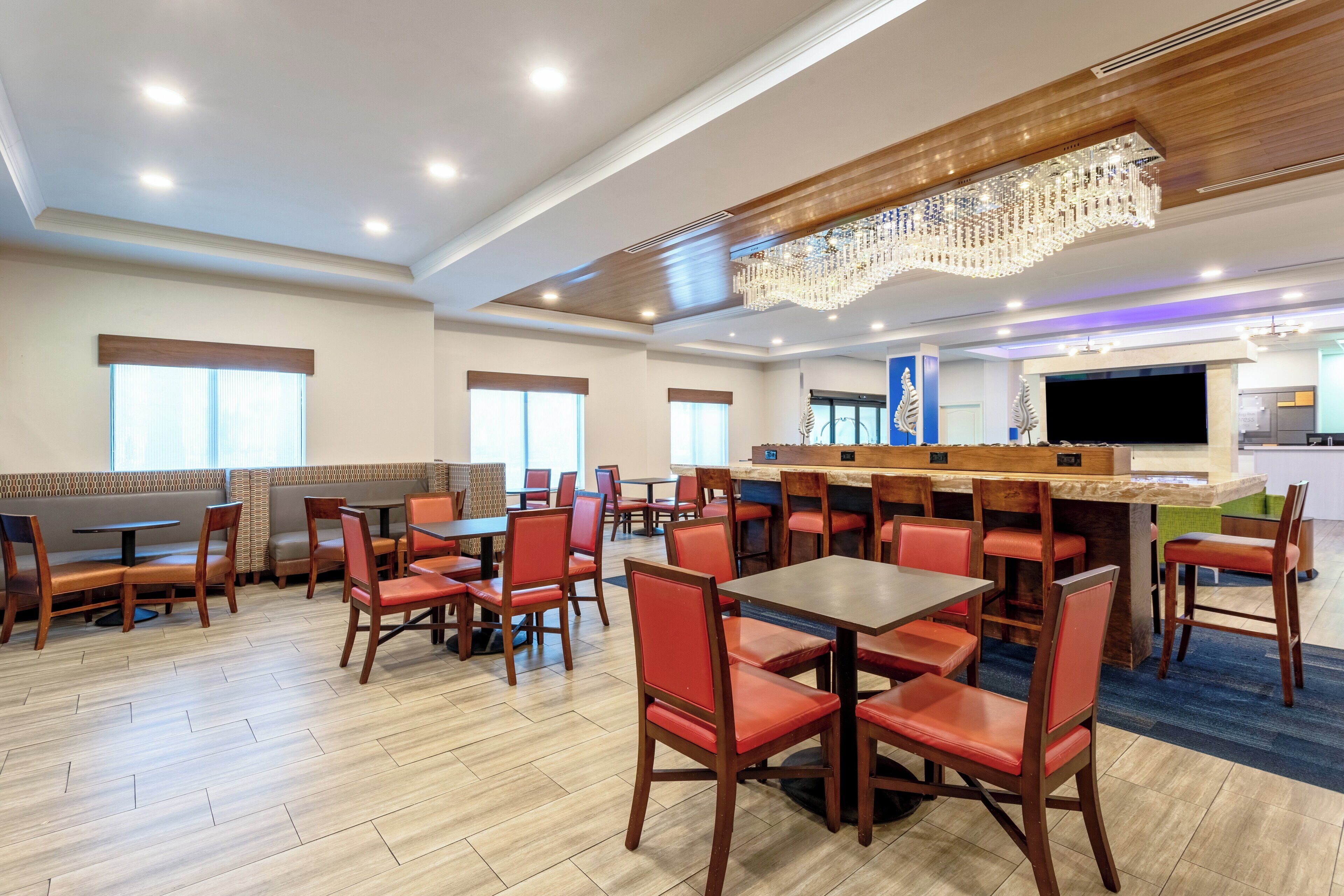 A spacious dining area with wooden tables and red chairs, a large chandelier, and a television screen.