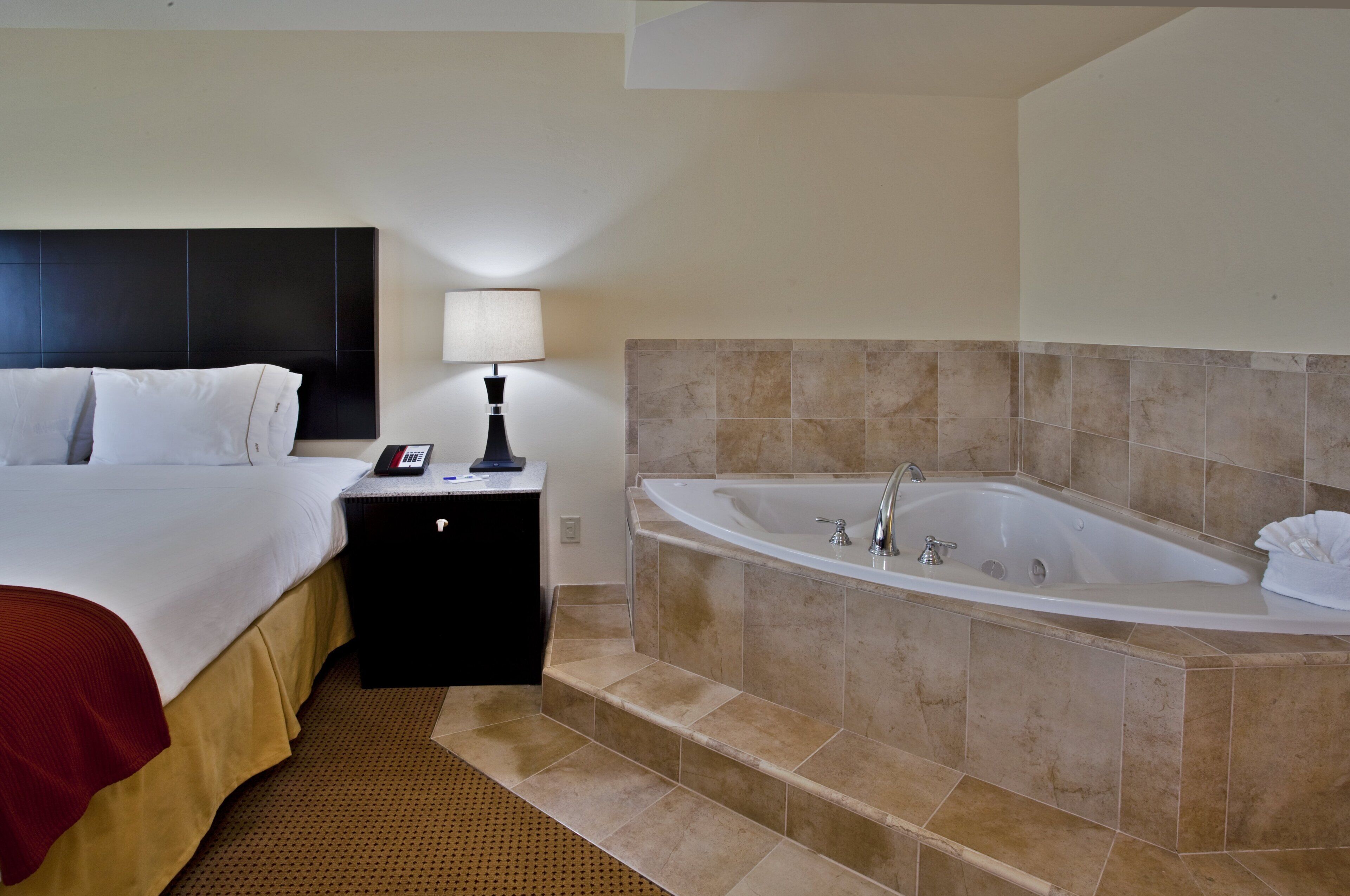 Una habitación de hotel con una cama, una mesita de noche con lámpara y una bañera.