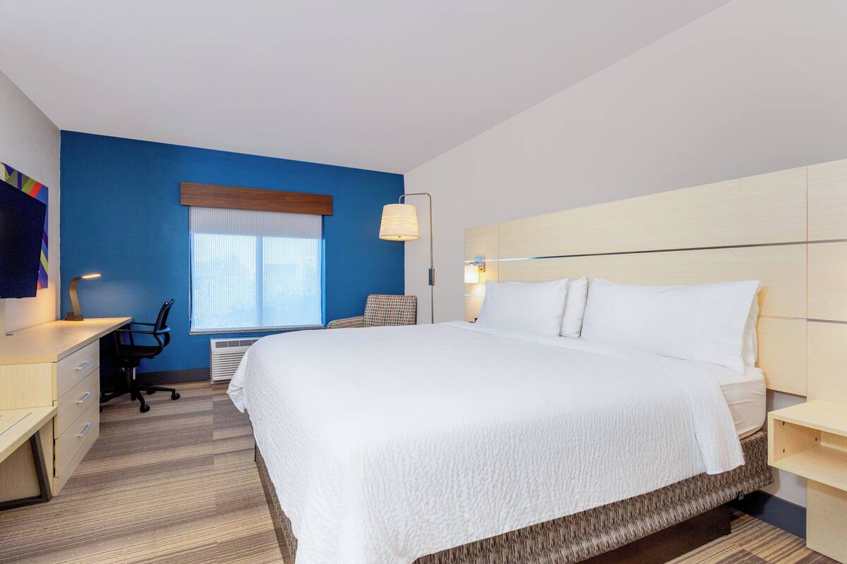 A hotel room with a large bed, a desk, a chair, and a window with blinds.