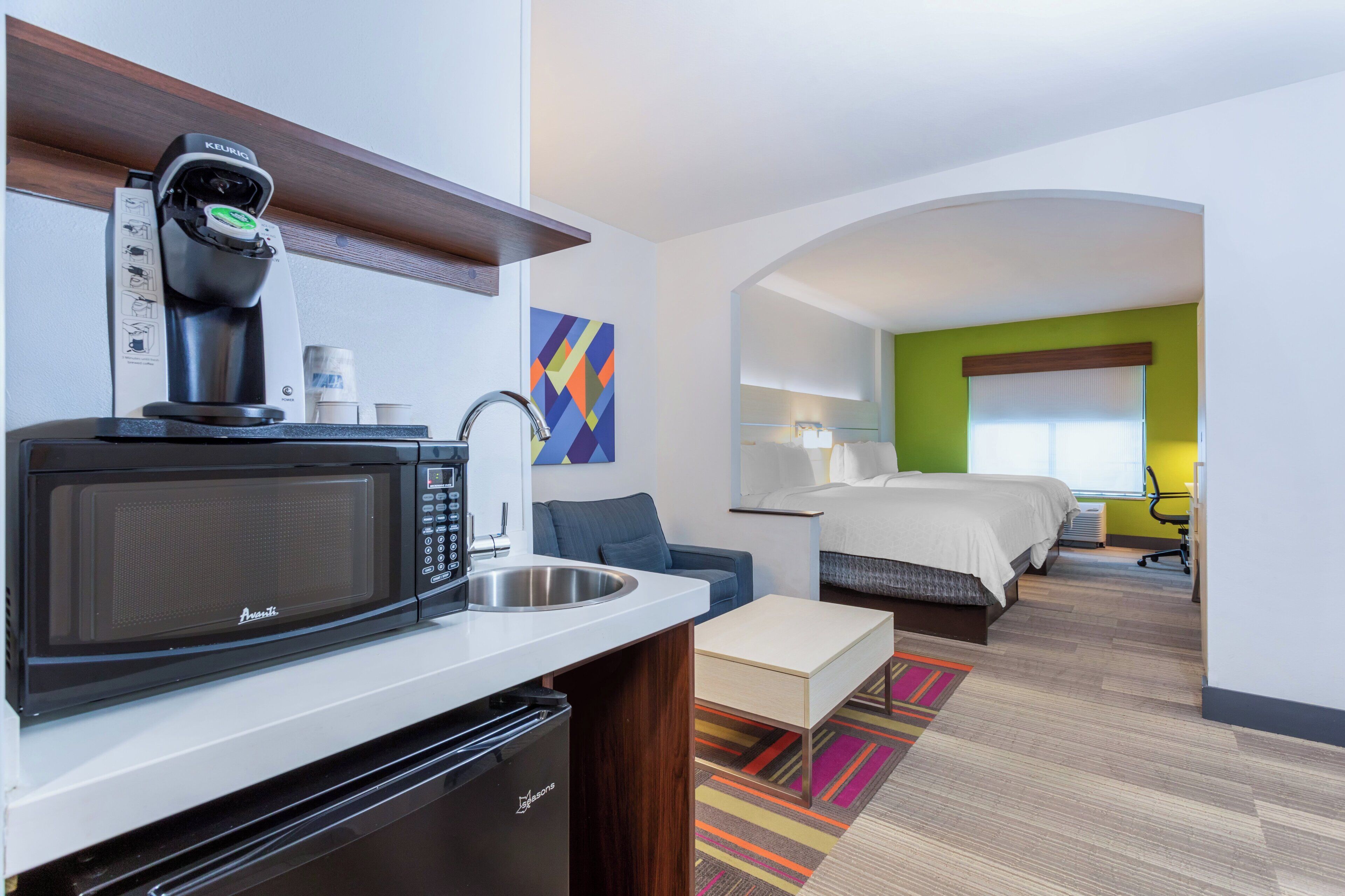 A compact hotel room with a kitchenette, a bed, a desk, and a view of the cityscape.