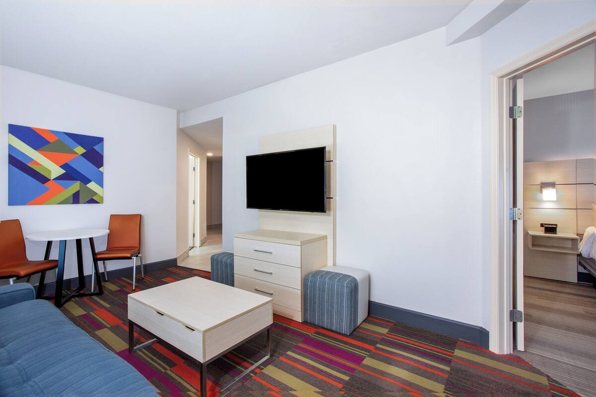 A modern hotel room with a flat-screen TV, a sofa, a small table, and a colorful striped carpet.