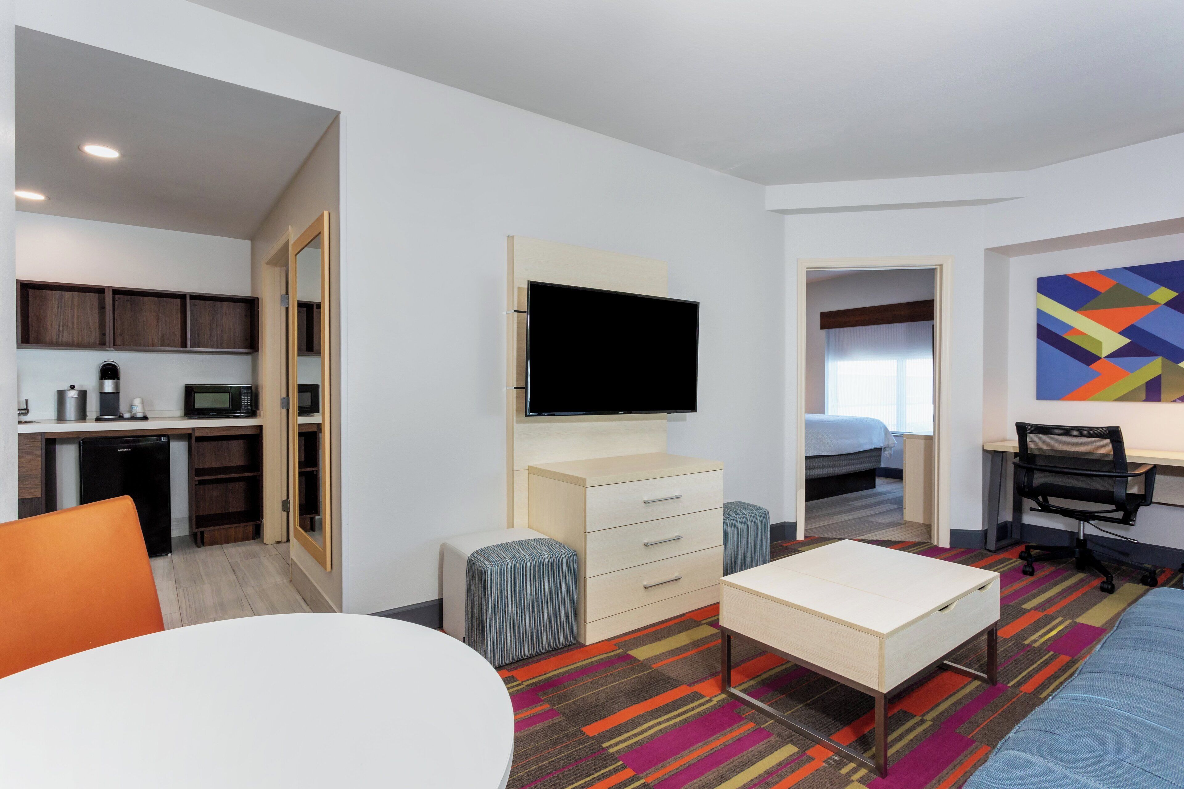 A hotel room with a kitchenette, a TV, a bed, a desk, and a colorful carpet.