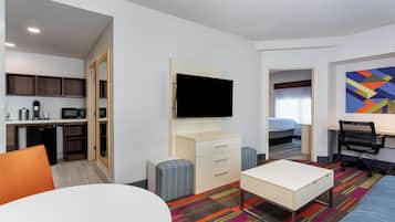 A hotel room with a kitchenette, a TV, a bed, a desk, and a colorful carpet.