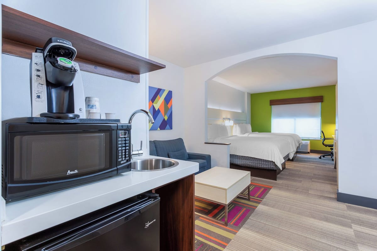 A compact hotel room with a kitchenette, a bed, a desk, and a view of the cityscape.
