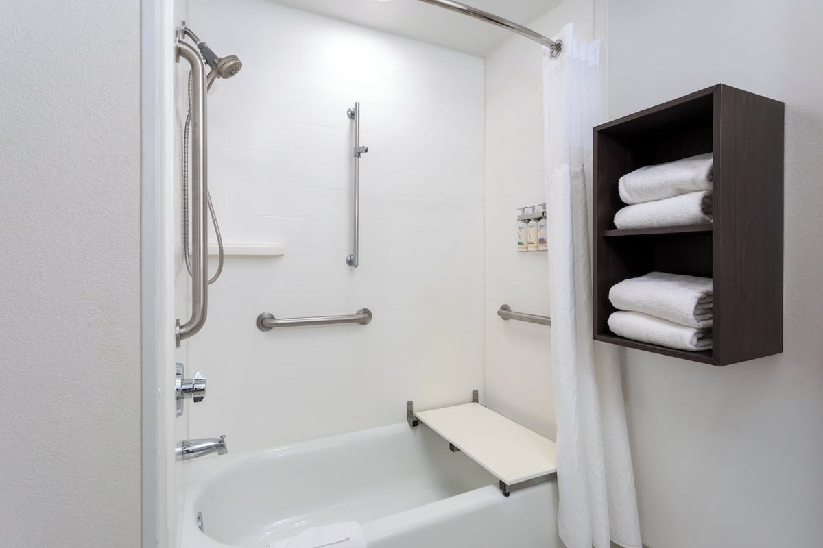 A bathroom with a white bathtub, grab bars, a shelf with towels, and a shower area.