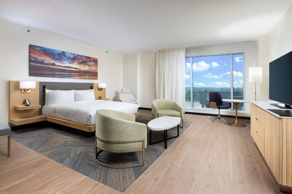 Ein Hotelzimmer mit einem Bett, zwei Sesseln, einem kleinen Tisch, einem Fernseher und einem großen Fenster mit Blick auf die Stadt.