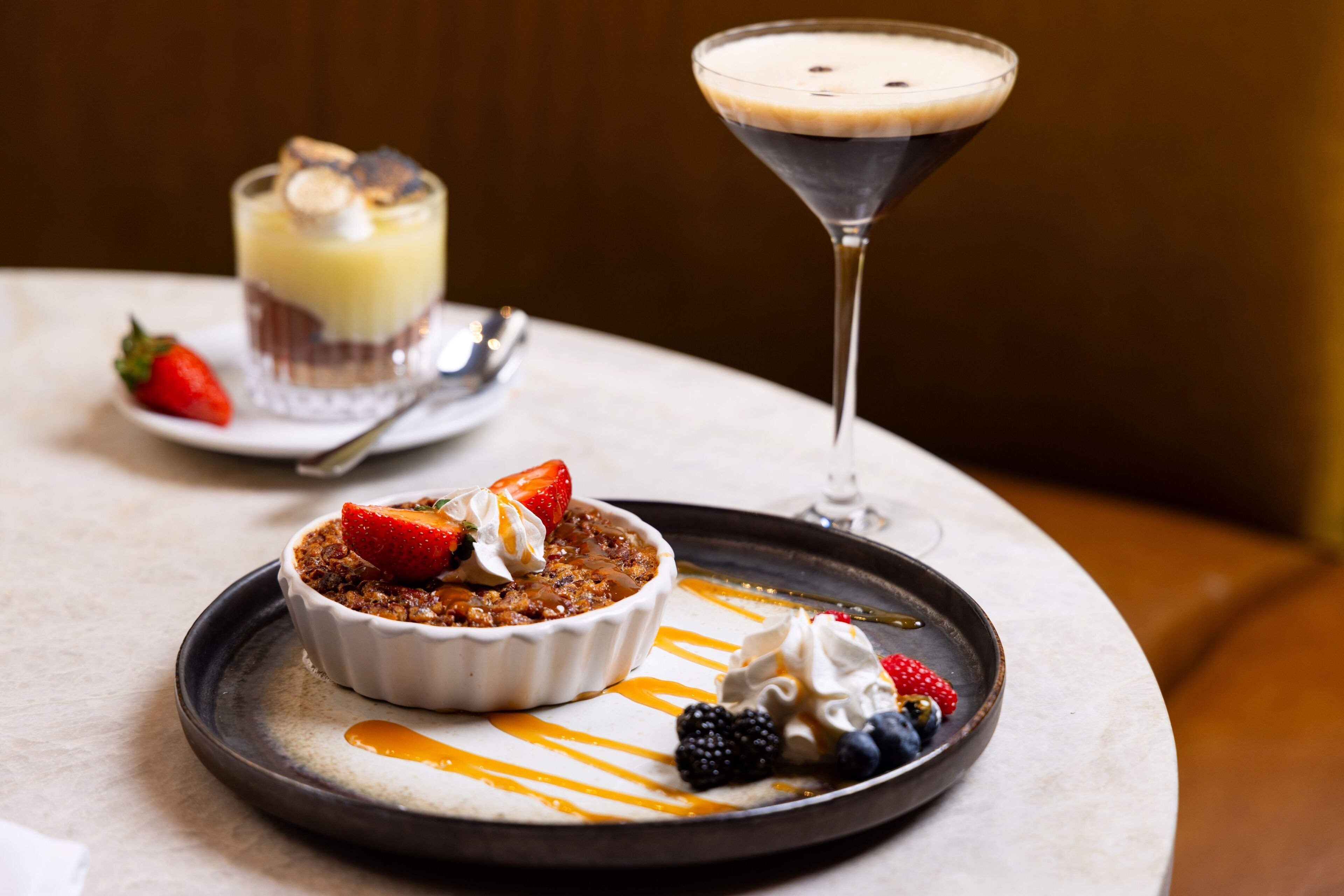 A dessert plate with a bowl of fruit crumble, a scoop of ice cream, and a martini glass with a dark-colored drink.