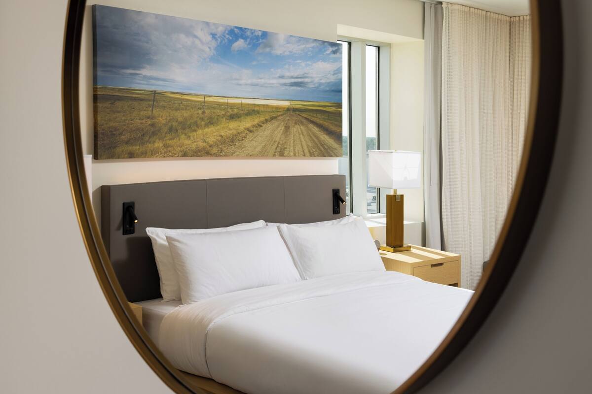 Ein Hotelzimmer mit Bett, Nachttisch, Lampe, einem großen Fenster und einem Landschaftsbild.