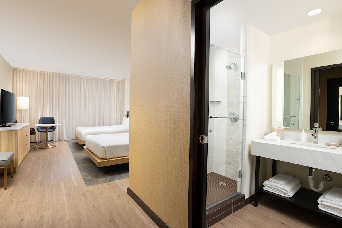 Ein Hotelzimmer mit Bett, Schreibtisch mit Stuhl, Fernseher, Badezimmer mit Waschbecken und Spiegel sowie einer Duschkabine.