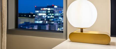A table lamp with a round white shade on a white table by a window with a city view.