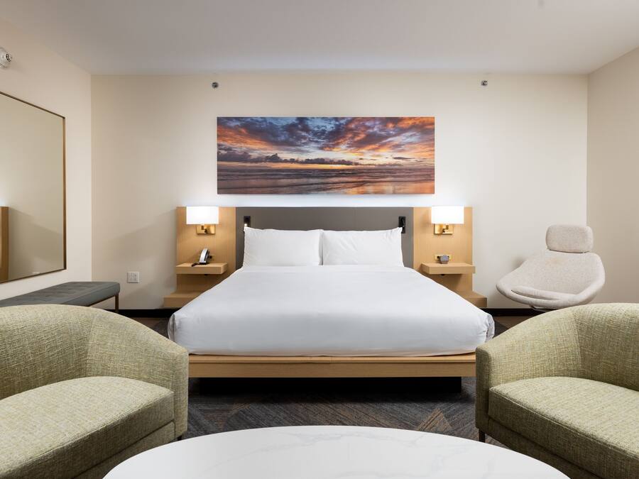 A hotel room with a large bed, two bedside tables with lamps, a chair, a round table, and a large painting of a sunset on the wall.